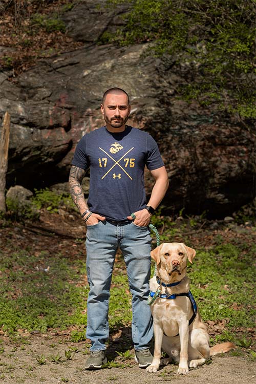 man standing in the forest with yellow lab in blue service vest sitting beside him