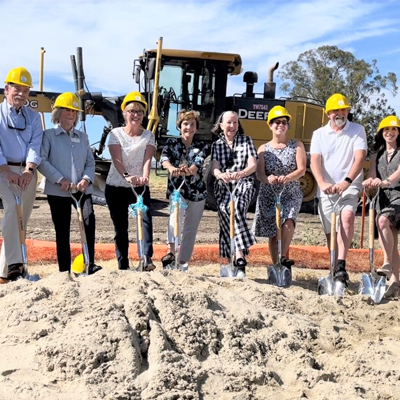 13 people with large shovels placed in the ground with a mound of sand and a bulldozer behind them