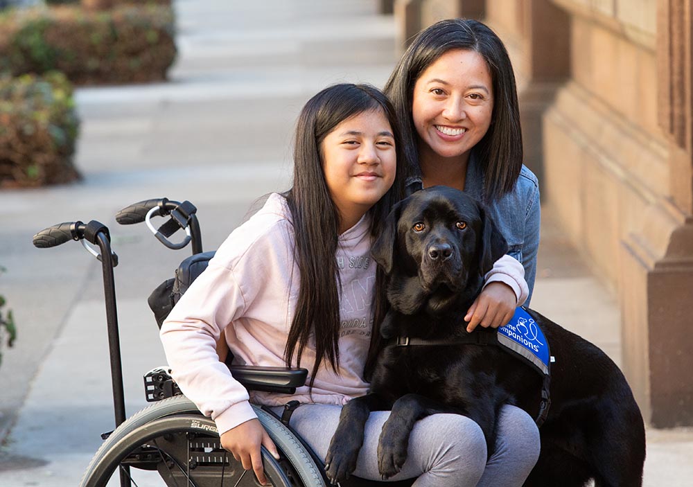 young girl in wheelchair smiles as black service dog lays across lap with her mother smiling behind them