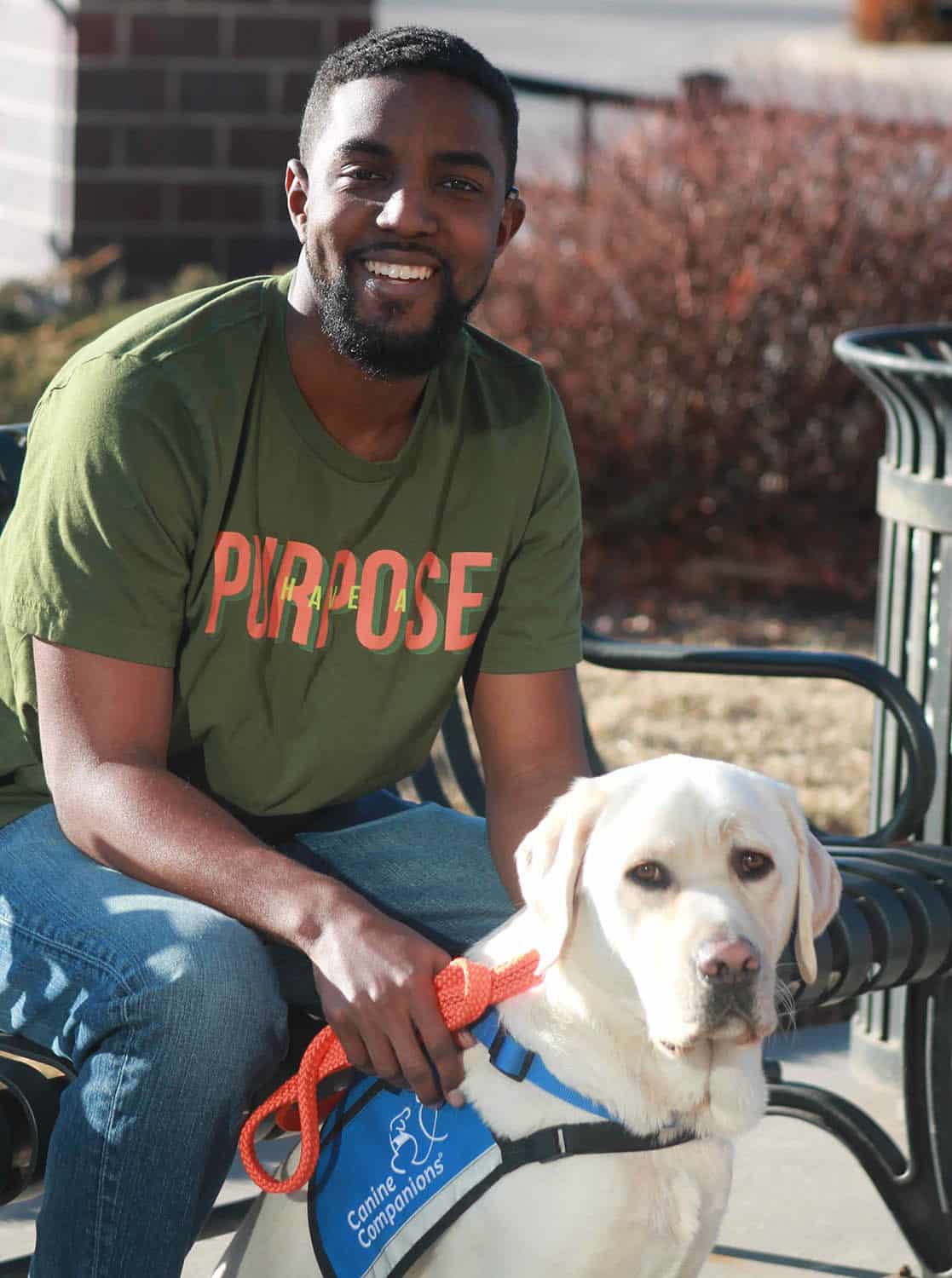 A smiling african american man holds the orange leash of his yellow lab service dog in blue vest