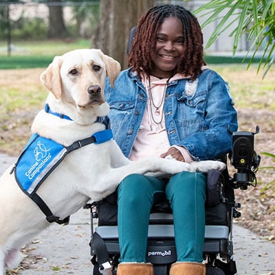 a woman sitting in a wheelchair smiling, as a Labrador wearing a blue vest with Canine Companions embroidery has its front legs on her lap