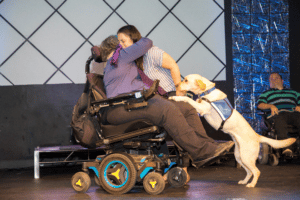 Woman in wheelchair hugging woman with yellow lab in blue vest on a stage