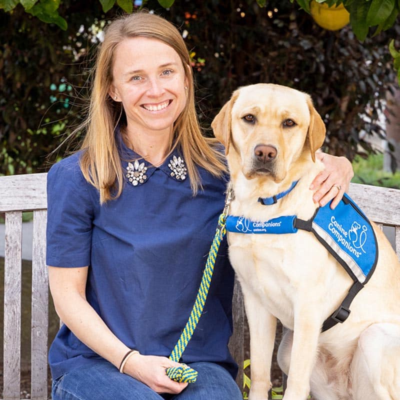 A smiling woman on a bench with her arm around a yellow lab in a blue service vest