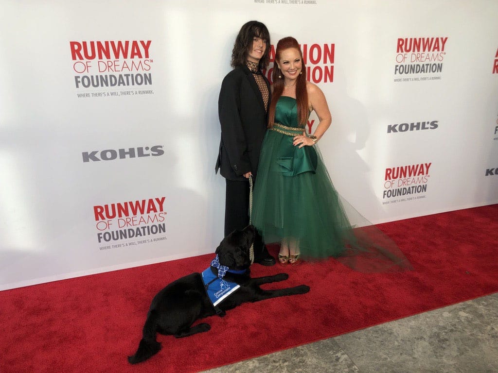 Two women on the red carpet at the Runway of Dreams Fashion Show with a black lab in blue service vest laying next to them