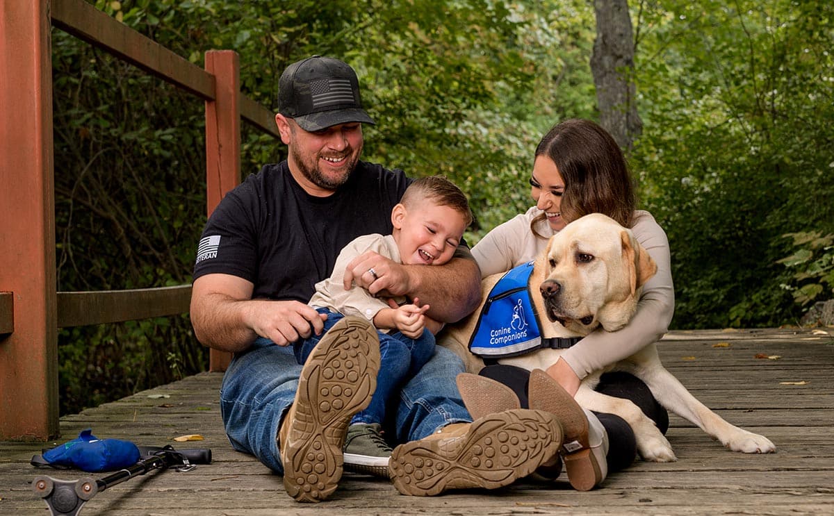 A smiling man in a military hat sits on the ground with his young son in his lap, his wife sitting next to him, and his yellow lab service dog laying in the middle.