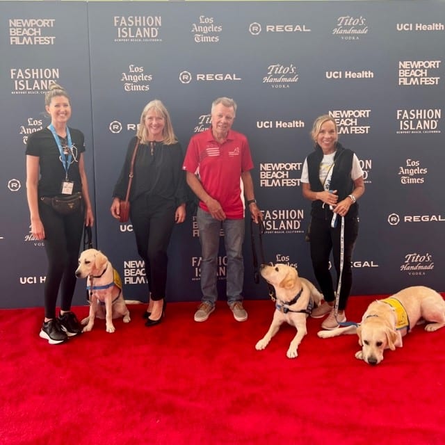 A group of people with yellow labs in yellow puppy vests on a red carpet at the newport beach film festival