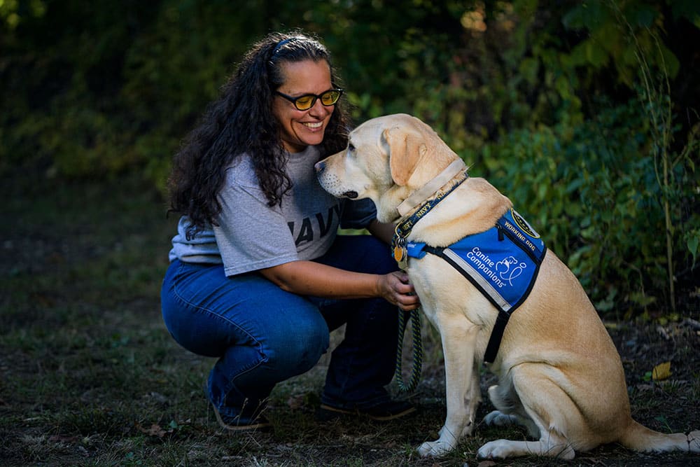 A woman bends down to pet her yellow lab service dog in a blue service vest.