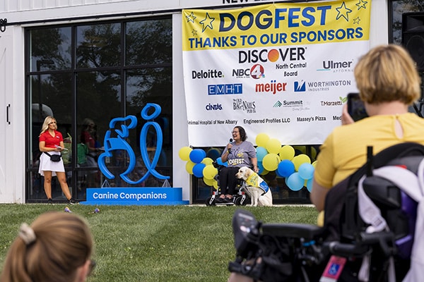 Woman in a wheelchair speaking on stage at the DogFest event with her service dog sitting next to her