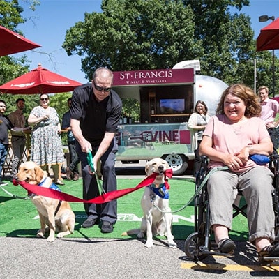 A group of adults and two Labradors holding a ribbon in mouth for a ribbon cutting ceremony; Airstream and parasols in the back