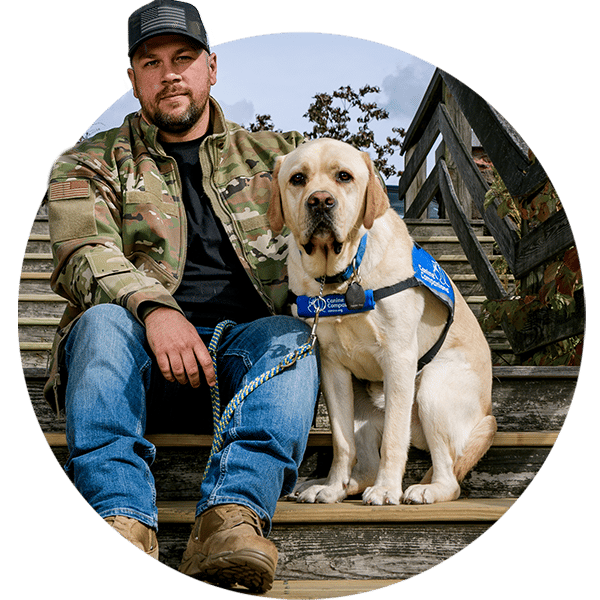 A man in camouflage sits on a stairway next to his yellow lab service dog