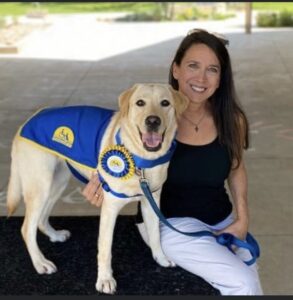 portrait of a smiling brunette woman next to a yellow lab in a blue service vest
