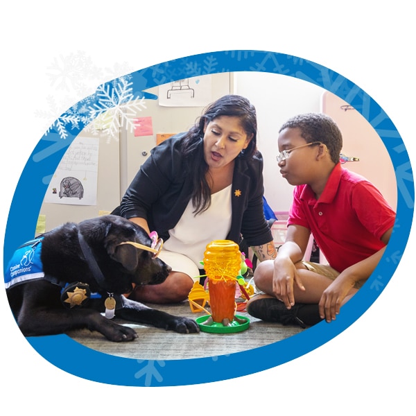 A blue circle with snowflakes around the image of a black lab facility dog playing a game with a young boy and the facility dog's handler