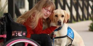 a young girl in a wheelchair hunching over hugging a Labrador sitting on a pavement outside