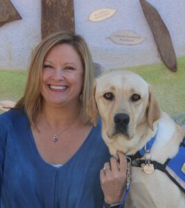 portrait of a smiling blond woman next to a yellow lab in a blue service vest