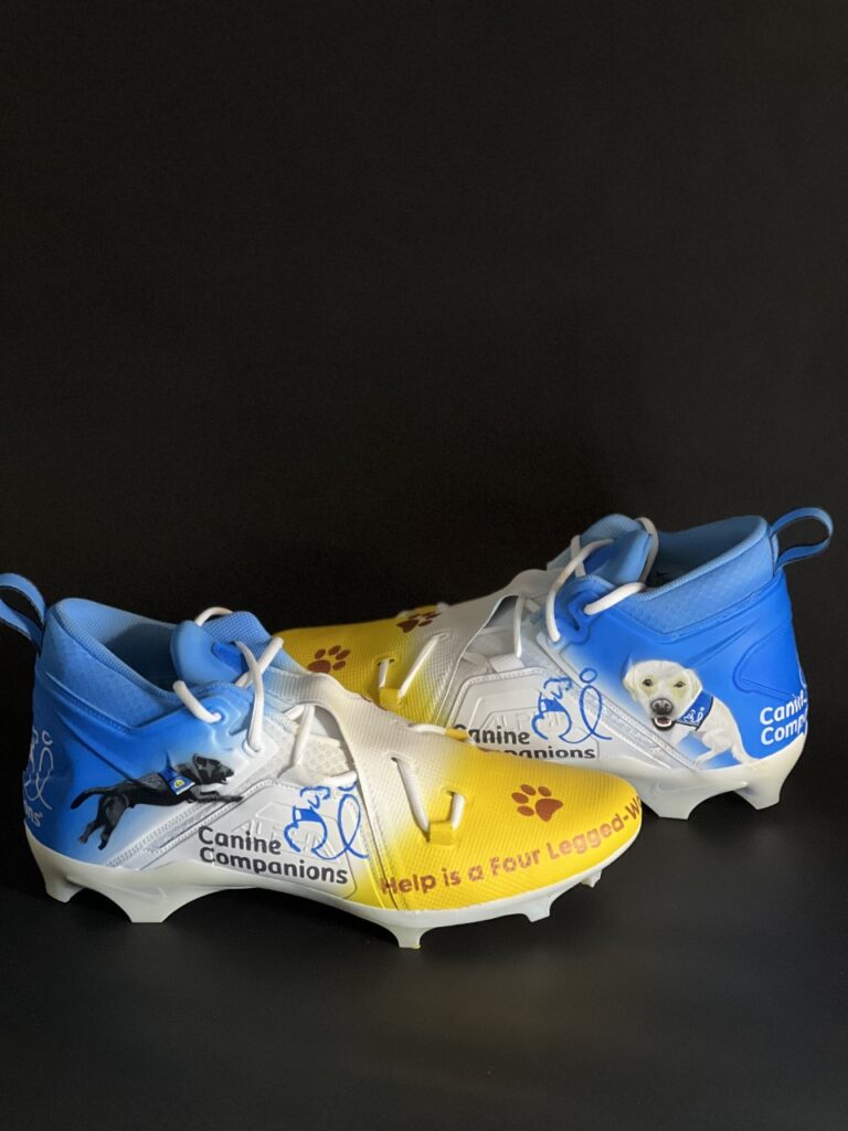 Yellow and blue cleats designed to represent Canine Companions feature a logo and two service dogs painted on the sides.