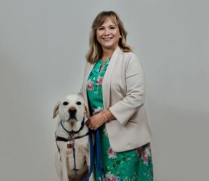 A smiling woman next to a yellow lab in a blue service vest