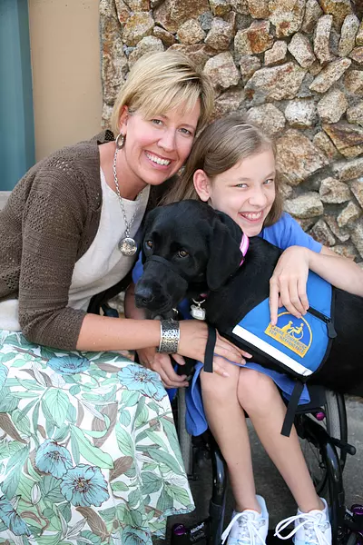 A young girl hugs her black lab service dog in a blue vest. The girls mother sits next to them hugging them both