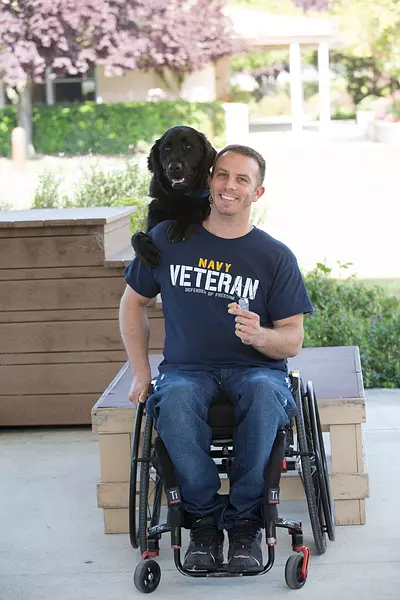 A man in a wheelchair smiles at the camera while a black lab service dog looks over his shoulder from behind