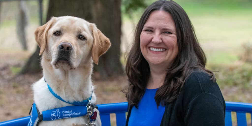 A smiling woman sits on a bench next to her yellow lab Facility Dog in a blue service vest