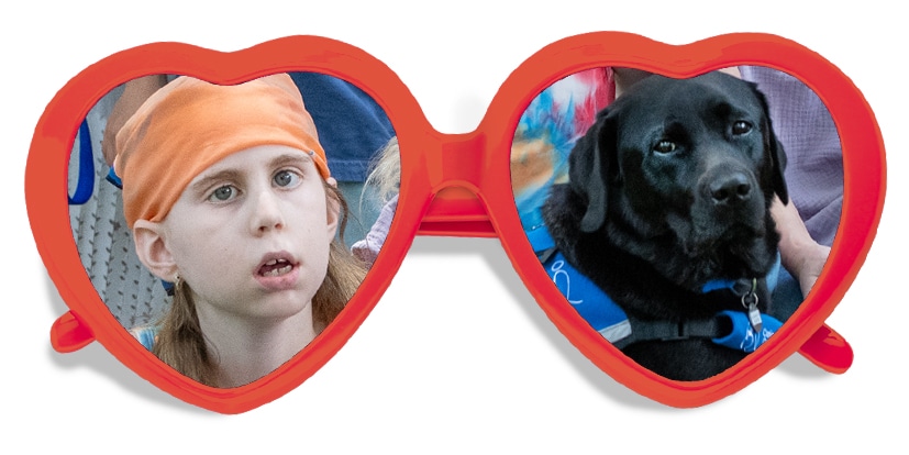 A red pair of heart shaped glasses with the image of a young girl and a black lab service dog in each lens.