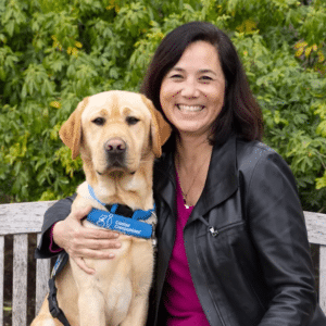 A smiling woman on a bench with her arm around a yellow lab in a blue service vest