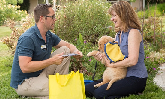 A man hands information to a woman holding a young puppy in a yellow puppy cape.