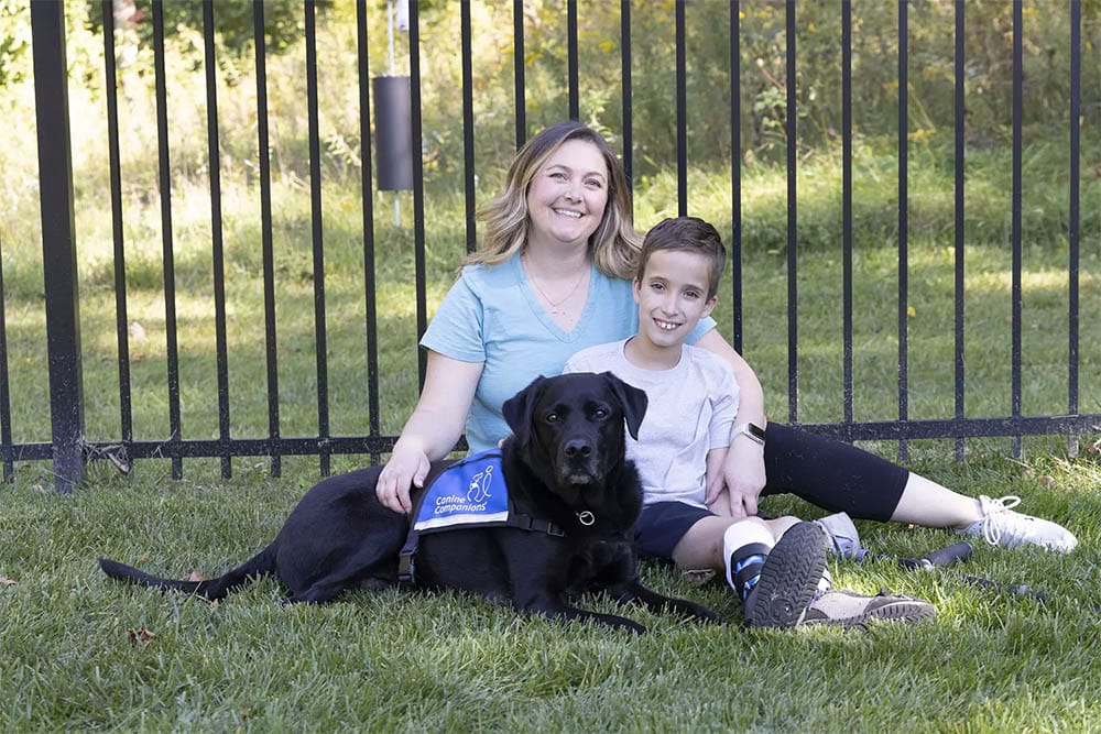 A young boy sits on the grass with his mother and his black lab service dog wearing a blue vest