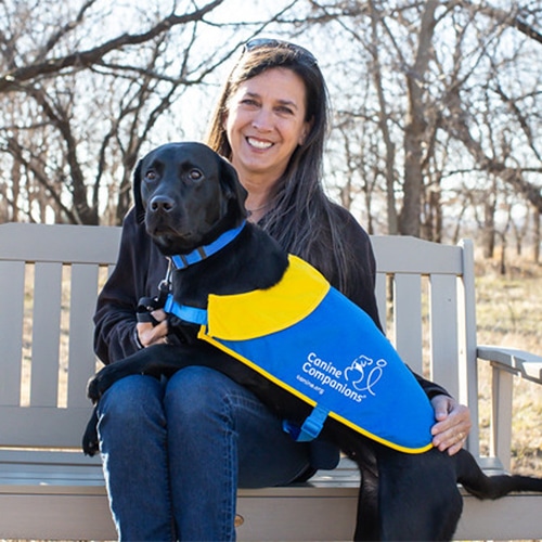 A smiling woman sitting on a bench outdoors with a black lab in a canine companions matriculation cape laying across her lap