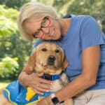 A smiling woman hugging a yellow lab puppy in a blue service cape