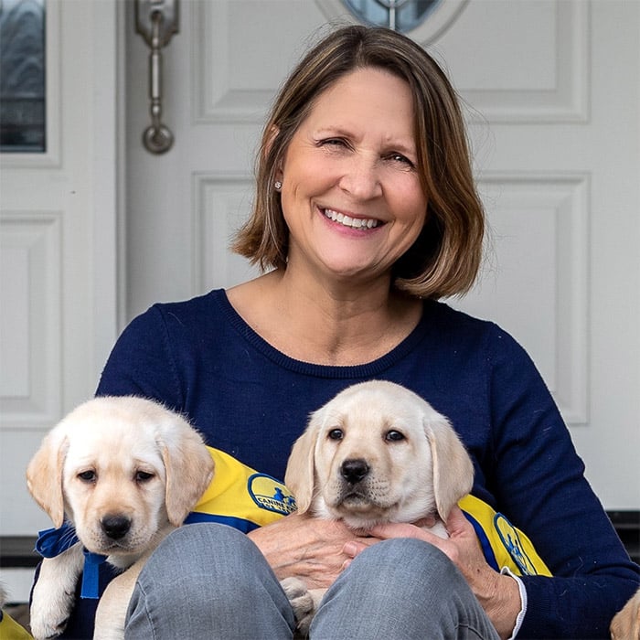 A smiling woman holding two puppies wearing yellow puppy capes