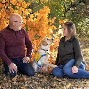 A man and woman sit with a yellow lab puppy in a patch of orange fall leaves. The puppy, which is wearing a yellow vest that reads “Canine Companions,” looks up at the woman.