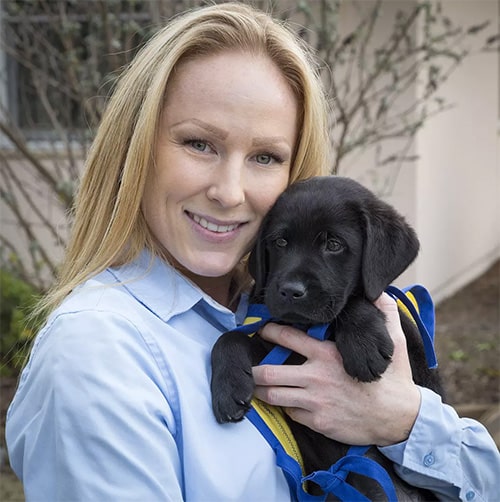 A smiling woman holds a black lab puppy in a yellow puppy cape