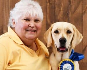 a smiling woman next to a yellow lab wearing a canine companions ribbon