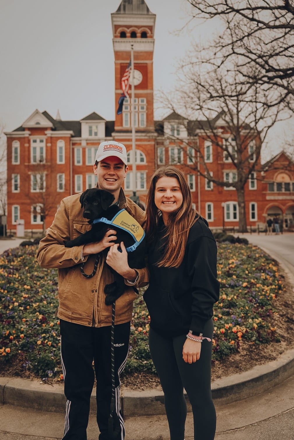 A young man holding a black lab puppy in a yellow puppy vest, standing on a college campus next to a young woman