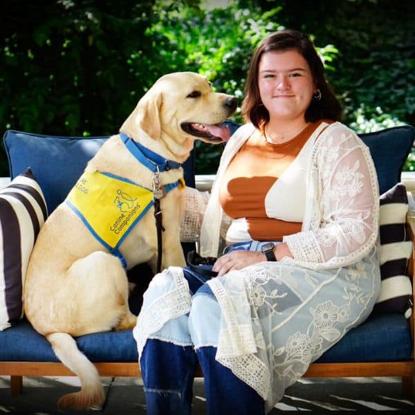 a young woman sitting with a Labrador puppy, who is wearing a yellow vest with Canine Companions written on it
