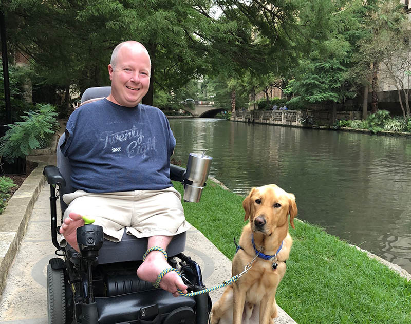 a man in a motorized wheelchair with a yellow lab service dog sitting beside him