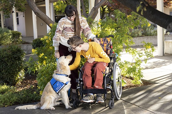a boy in a wheelchair bends down to pet his yellow lab service dog wearing a blue service vest. His mother stand behind him.