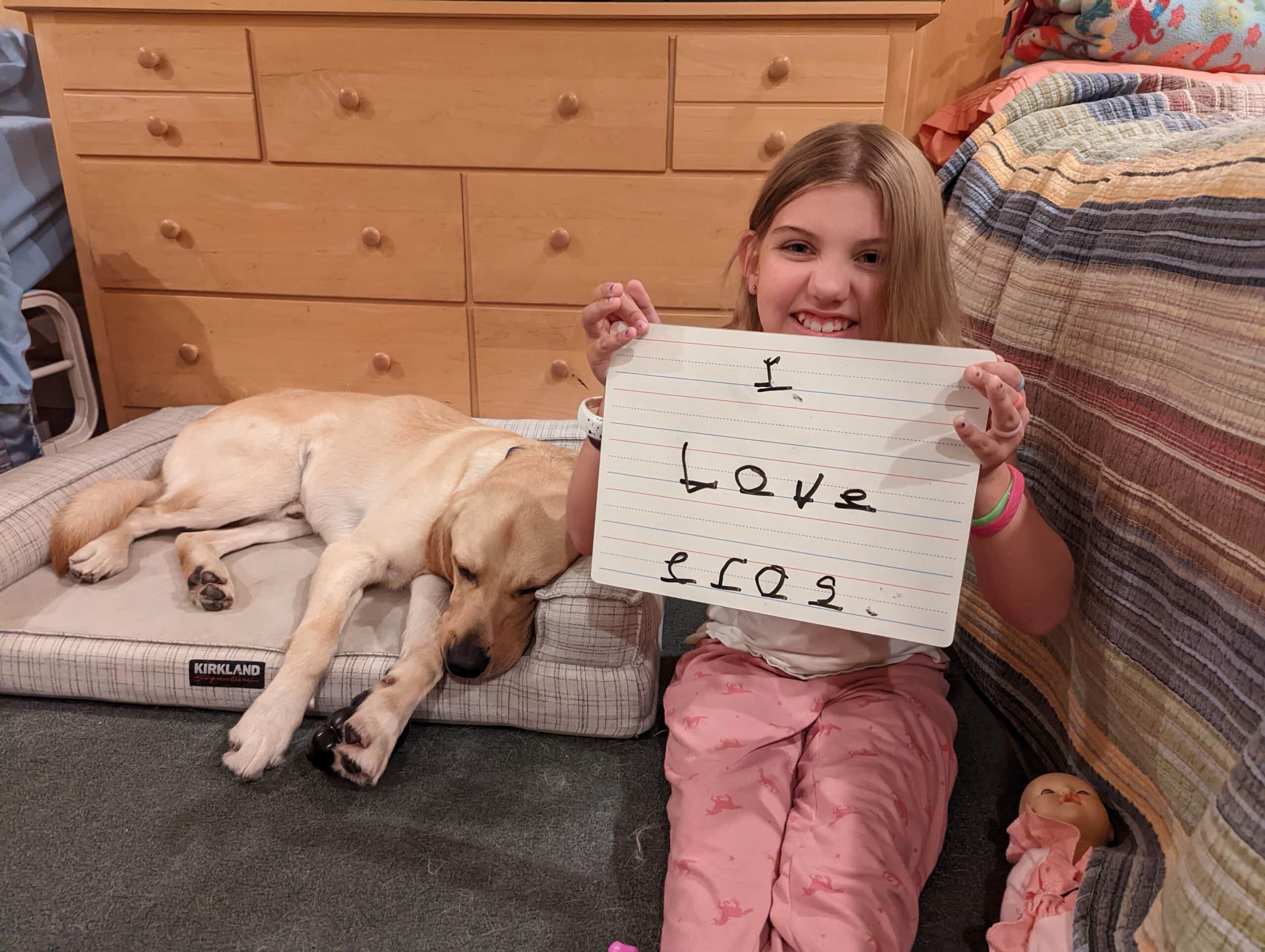 A smiling young girl with a sign that says I love Eros sitting next to her sleeping yellow lab service dog