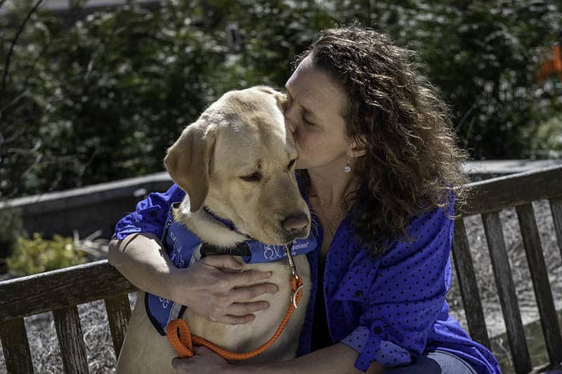 a woman kissing a yellow lab in a blue service dog vest