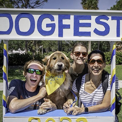 three smiling adults with a golden retriever in a yellow dogfest bandana in a photo booth that says DOGFEST