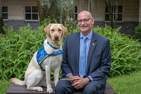A smiling man in a suit sits next to a yellow lab service dog in a blue service vest