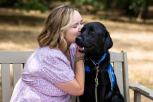A woman sitting on a bench outdoors kissing her black Labrador/Golden retriever cross service dog wearing a blue vest