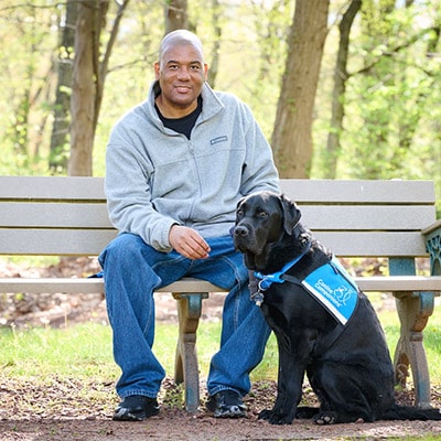 a man sitting on a bench in the woods, with a Labrador sitting next to him, wearing a blue vest that reads, Canine Companions