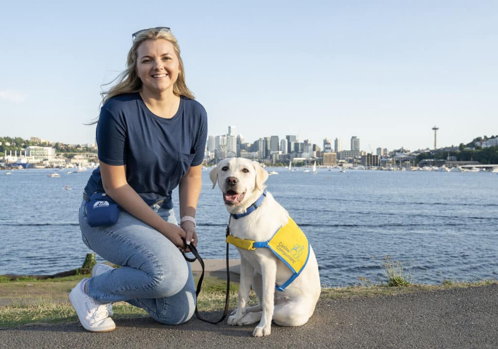 a smiling young woman with a yellow lab in a yellow puppy vest on the Seattle bayfront