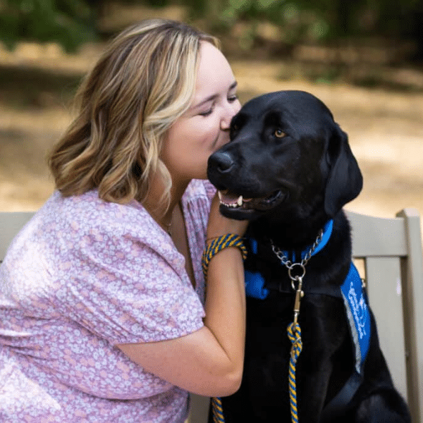 a woman sitting on a bench kissing a Labrador who is wearing a blue vest and a leash attached