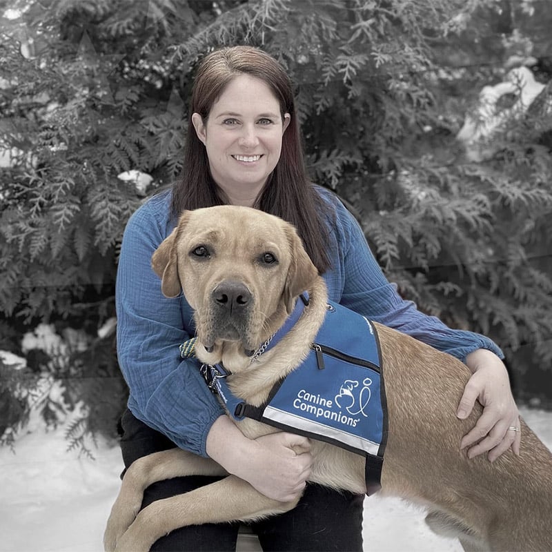 A smiling woman with a yellow lab in a blue service vest across her lap