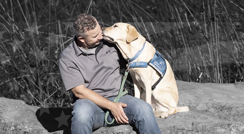 A man looking at his yellow lab wearing a blue service vest who is licking the mans face.