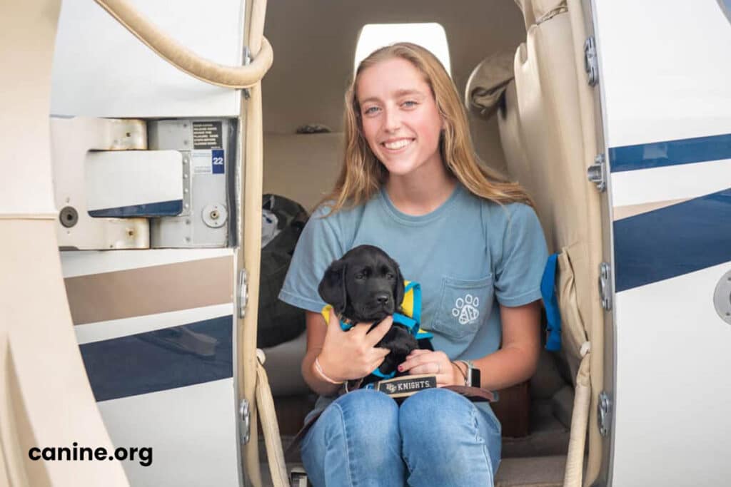 A smiling girl sitting in the doorway of a small aircraft holding a black lab puppy