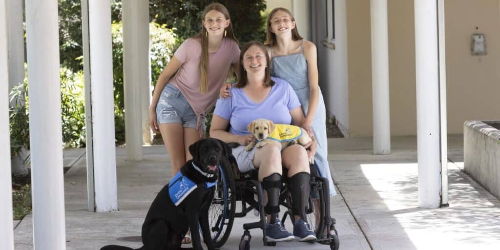 A smiling woman in a wheelchair holds a yellow lab puppy in a yellow puppy cape in her lap. She is surrounded by two smiling girls and a black lab in a service vest