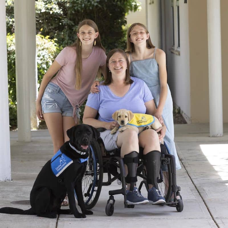 A smiling woman in a wheelchair holds a yellow lab puppy in a yellow puppy cape in her lap. She is surrounded by two smiling girls and a black lab in a service vest